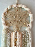 This dreamcatcher has an antique sand-colored star-shaped vintage doily. Stitched in the center is a circle of brown and white shell beads. Trailing below are cream and white yarns rough spun and smooth velveteen in texture. Handmade by Sarah Moody. 