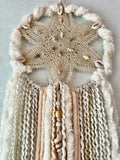 Close up of - This dreamcatcher has an antique sand-colored star-shaped vintage doily. Stitched in the center is a circle of brown and white shell beads. Trailing below are cream and white yarns rough spun and smooth velveteen in texture. Handmade by Sarah Moody.  Dreamcatcher measures: 28" in length and 8.5" wide.